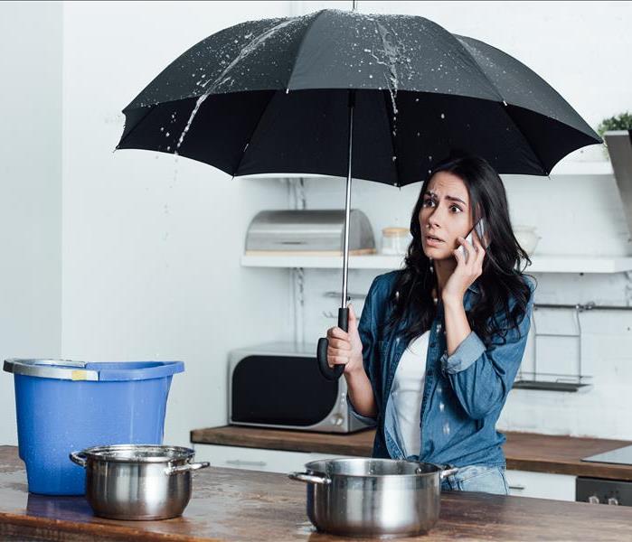 Shocked woman with umbrella dealing with water damage in kitchen and talking on smartphone